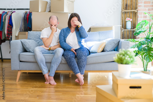 Young couple sitting on the sofa arround cardboard boxes moving to a new house tired rubbing nose and eyes feeling fatigue and headache. Stress and frustration concept.