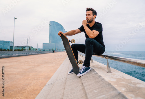 Skateboarder sits with skateboard on the modern city sea terrace. © Igor Kardasov