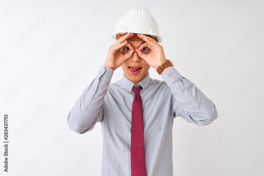 Chinese architect man wearing tie and helmet standing over isolated white background doing ok gesture like binoculars sticking tongue out, eyes looking through fingers. Crazy expression. Chinese architect man wearing tie and helmet standing over isolated white background doing ok gesture like binoculars sticking tongue out, eyes looking through fingers. Crazy expression.