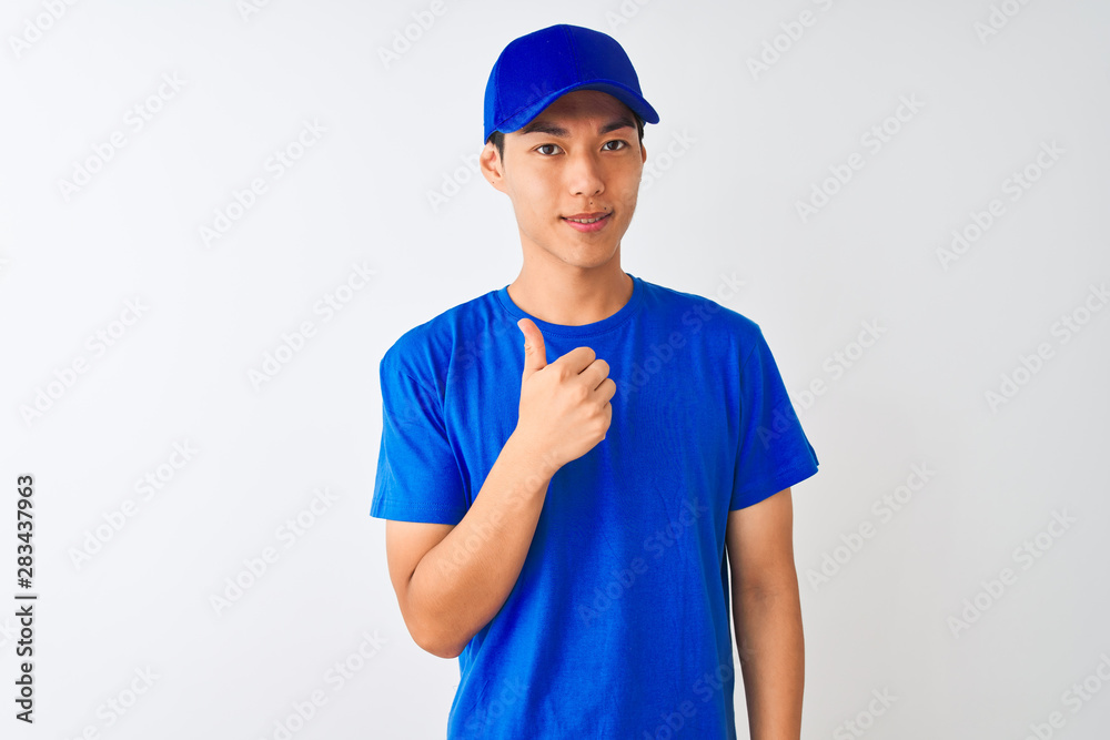 Chinese deliveryman wearing blue t-shirt and cap standing over isolated white background doing happy thumbs up gesture with hand. Approving expression looking at the camera showing success.