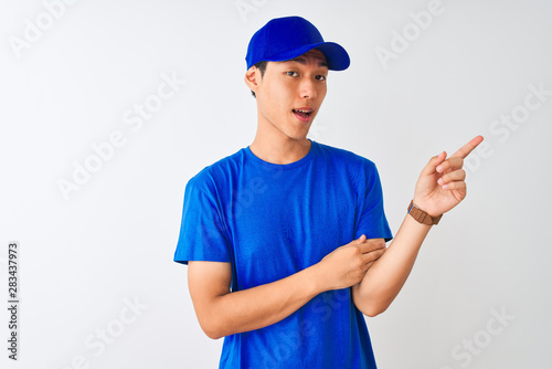 Chinese deliveryman wearing blue t-shirt and cap standing over isolated white background with a big smile on face, pointing with hand and finger to the side looking at the camera.