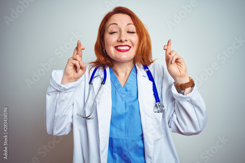 Young redhead doctor woman using stethoscope over white isolated background gesturing finger crossed smiling with hope and eyes closed. Luck and superstitious concept.