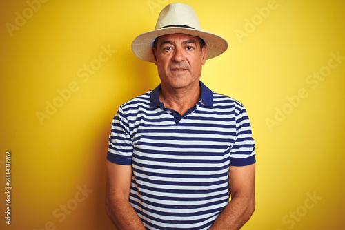 Handsome middle age man wearing striped polo and hat over isolated yellow background with serious expression on face. Simple and natural looking at the camera.