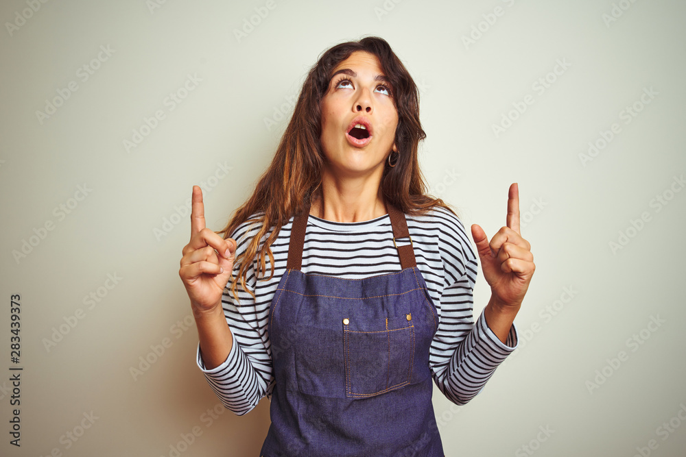 Young beautiful chef woman wearing apron cooking over white isolated background amazed and surprised looking up and pointing with fingers and raised arms. Young beautiful chef woman wearing apron cooking over white isolated background amazed and surprised looking up and pointing with fingers and raised arms.