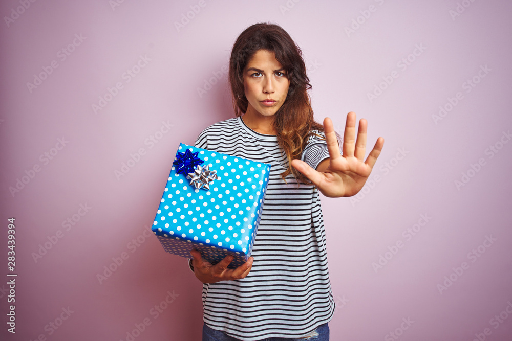 Young beautiful woman holding birthday gift over pink isolated background with open hand doing stop sign with serious and confident expression, defense gesture Young beautiful woman holding birthday gift over pink isolated background with open hand doing stop sign with serious and confident expression, defense gesture