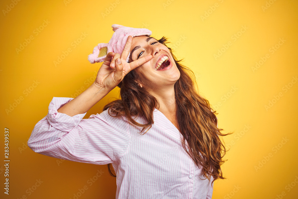 Young woman wearing pajama and sleep mask standing over yellow isolated background Doing peace symbol with fingers over face, smiling cheerful showing victory Young woman wearing pajama and sleep mask standing over yellow isolated background Doing peace symbol with fingers over face, smiling cheerful showing victory