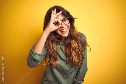Young beautiful woman wearing green shirt and glasses over yelllow isolated background doing ok gesture with hand smiling, eye looking through fingers with happy face. photo