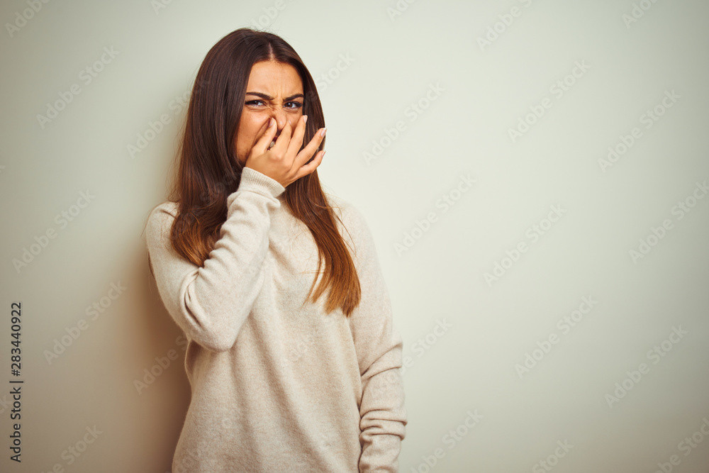 Young beautiful woman wearing winter turtleneck sweater over isolated white background smelling something stinky and disgusting, intolerable smell, holding breath with fingers on nose. Bad smells 