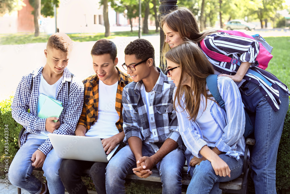 Young students with laptop sitting on bench outdoors