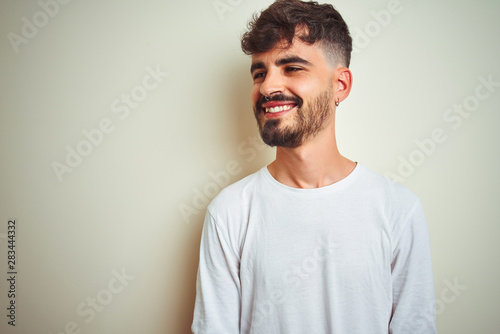 Young man with tattoo wearing t-shirt standing over isolated white background looking away to side with smile on face, natural expression. Laughing confident.