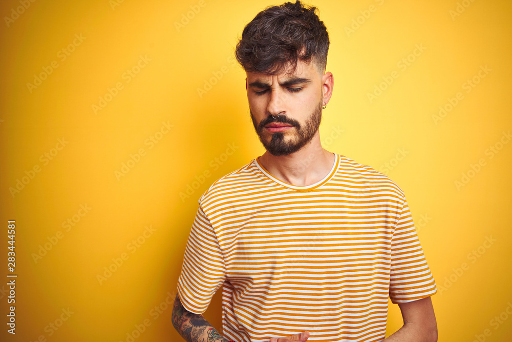 Young man with tattoo wearing striped t-shirt standing over isolated yellow background with hand on stomach because indigestion, painful illness feeling unwell. Ache concept. Young man with tattoo wearing striped t-shirt standing over isolated yellow background with hand on stomach because indigestion, painful illness feeling unwell. Ache concept.