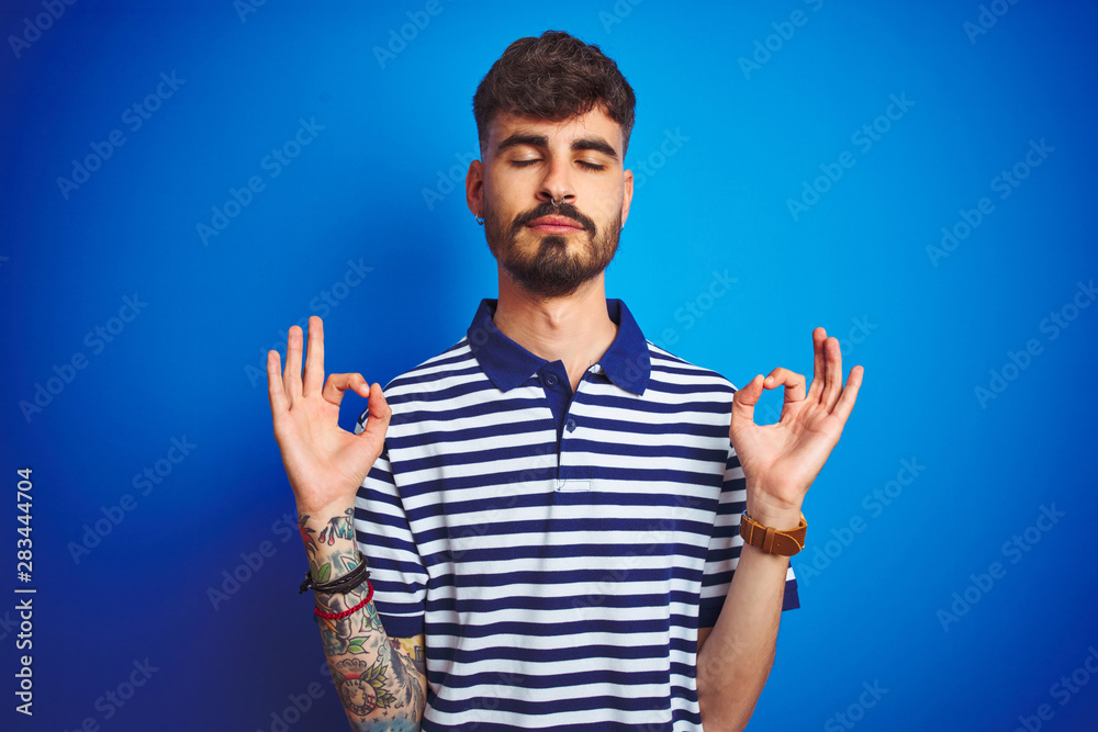 Fototapeta premium Young man with tattoo wearing striped polo standing over isolated blue background relax and smiling with eyes closed doing meditation gesture with fingers. Yoga concept.