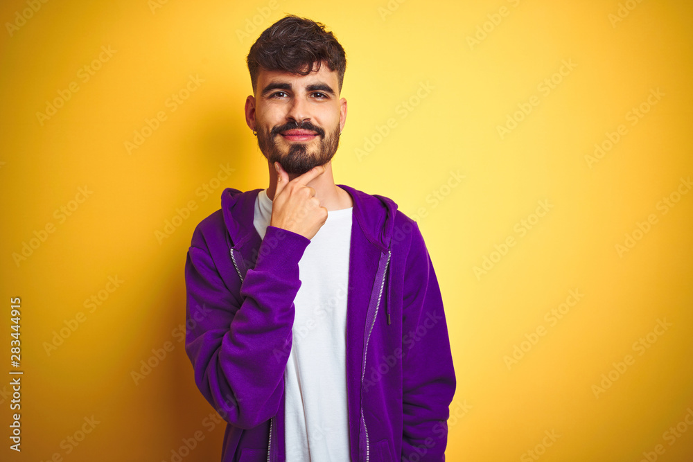 Young man with tattoo wearing sport purple sweatshirt over isolated yellow background looking confident at the camera with smile with crossed arms and hand raised on chin. Thinking positive. Young man with tattoo wearing sport purple sweatshirt over isolated yellow background looking confident at the camera with smile with crossed arms and hand raised on chin. Thinking positive.
