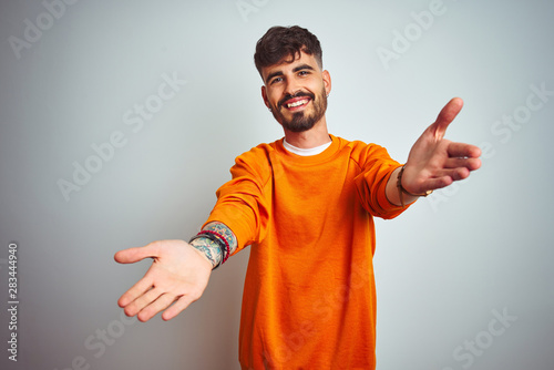 Photography Young man with tattoo wearing orange sweater standing over isolated white background looking at the camera smiling with open arms for hug