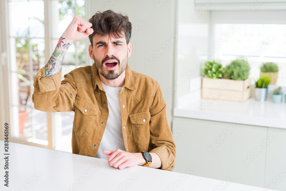 Young man wearing casual jacket sitting on white table angry and mad raising fist frustrated and furious while shouting with anger. Rage and aggressive concept.