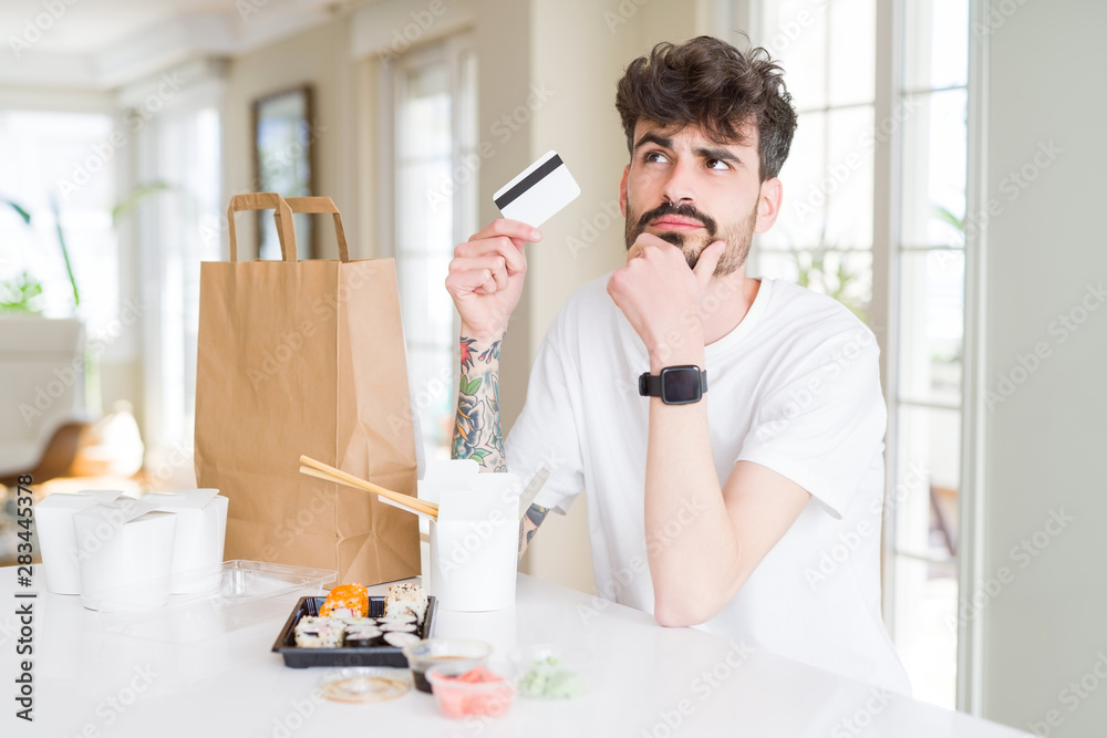 Young man eating asian sushi from home delivery using credit card as payment serious face thinking about question, very confused idea Young man eating asian sushi from home delivery using credit card as payment serious face thinking about question, very confused idea