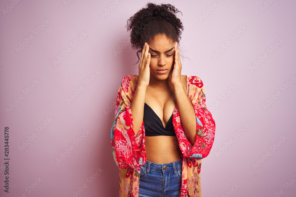American woman on holiday wearing bikini and colorful dress over isolated pink background with hand on head for pain in head because stress. Suffering migraine. American woman on holiday wearing bikini and colorful dress over isolated pink background with hand on head for pain in head because stress. Suffering migraine.