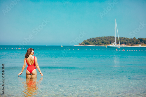 sexy woman in red swimsuit standing in sea water. coastline yacht on background