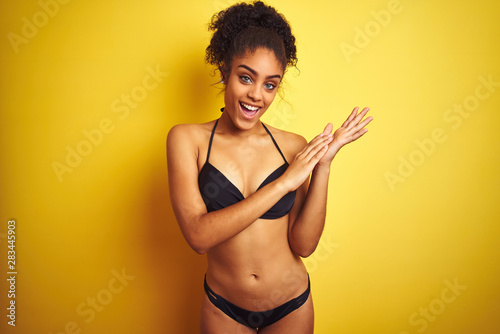 African american woman on vacation wearing bikini standing over isolated yellow background clapping and applauding happy and joyful, smiling proud hands together