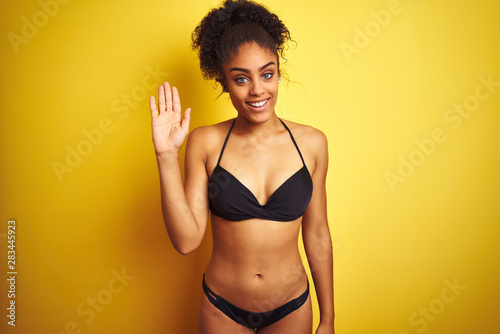 African american woman on vacation wearing bikini standing over isolated yellow background Waiving saying hello happy and smiling, friendly welcome gesture © Krakenimages.com