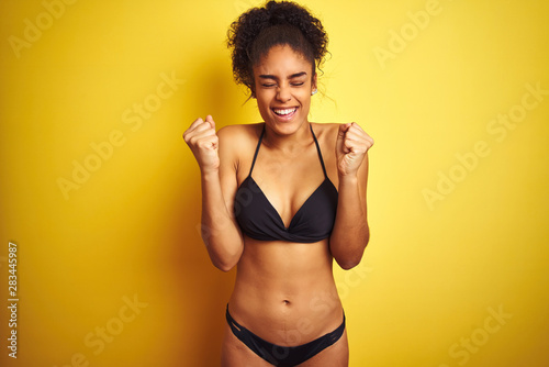 African american woman on vacation wearing bikini standing over isolated yellow background excited for success with arms raised and eyes closed celebrating victory smiling. Winner concept.