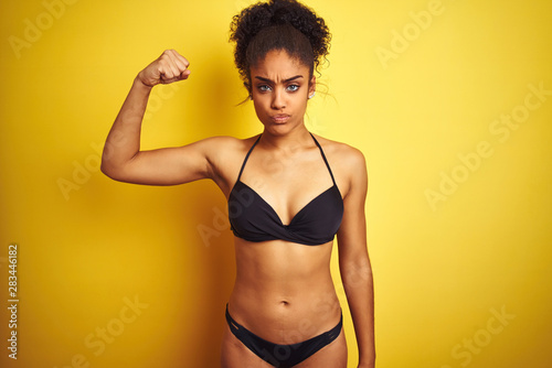 African american woman on vacation wearing bikini standing over isolated yellow background Strong person showing arm muscle, confident and proud of power © Krakenimages.com
