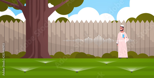arab man using mobile app controlling irrigation grass watering system by smart speaker voice recognition activated digital assistant concept backyard with green lawn horizontal full length