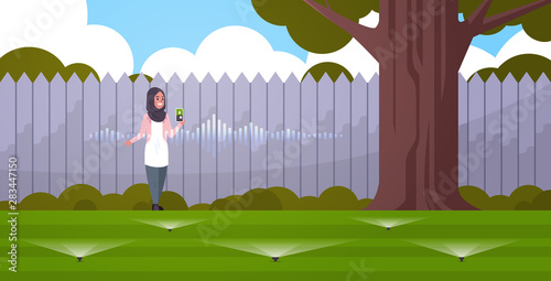 arab woman using mobile app controlling irrigation grass watering system by smart speaker voice recognition activated assistant concept backyard with green lawn horizontal full length