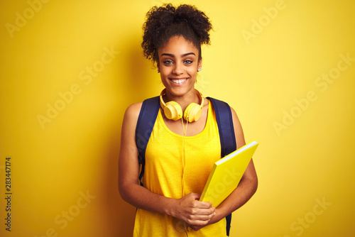 Afro woman using backpack and headphones holding notebook over isolated yellow background with a happy face standing and smiling with a confident smile showing teeth