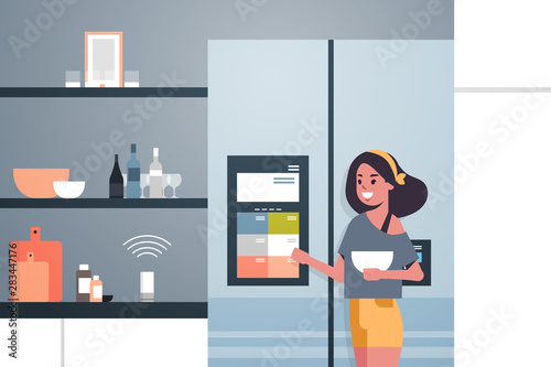 woman touching refrigerator screen with smart speaker voice recognition activated digital assistant concept modern kitchen interior flat horizontal portrait
