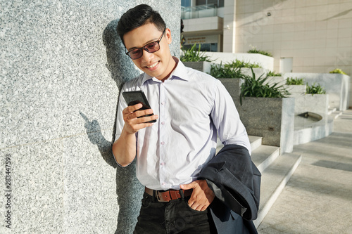 Happy young Vietnamese entrepreneur leaning on wall and checking social media in his smartphone