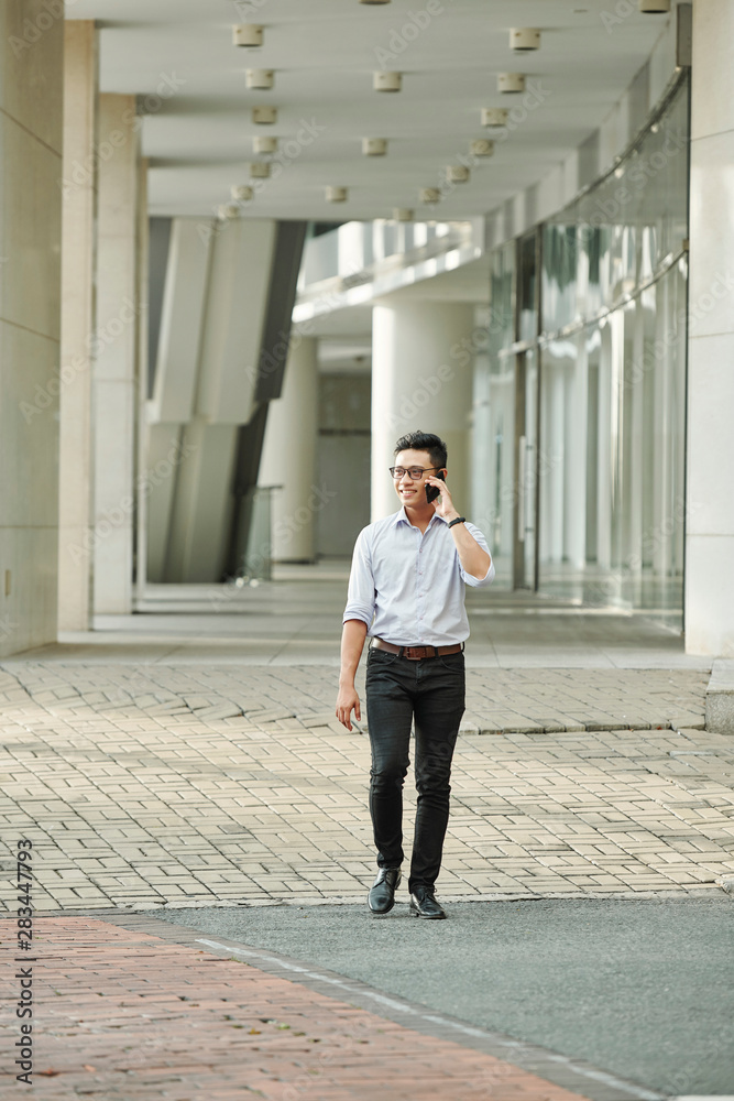 Confident smiling Vietnamese businessman in glasses walking outdoors and calling on phone