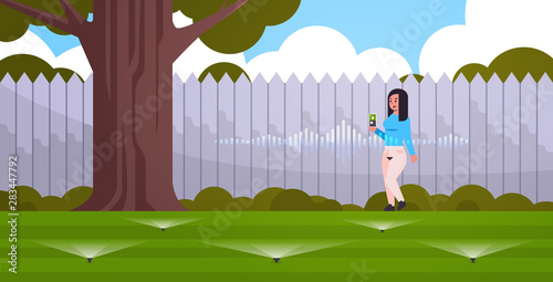 woman using mobile app controlling irrigation grass watering system by smart speaker voice recognition activated digital assistant concept modern backyard with green lawn horizontal full length