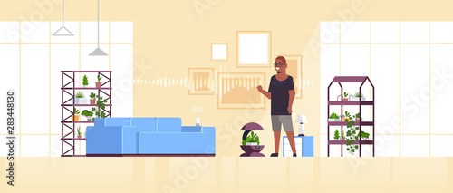 african american man using auto watering system controlled by smart speaker voice recognition activated digital assistant smart home concept living room interior full length horizontal