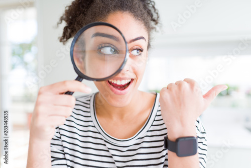 Young african american woman looking through magnifying glass pointing and showing with thumb up to the side with happy face smiling