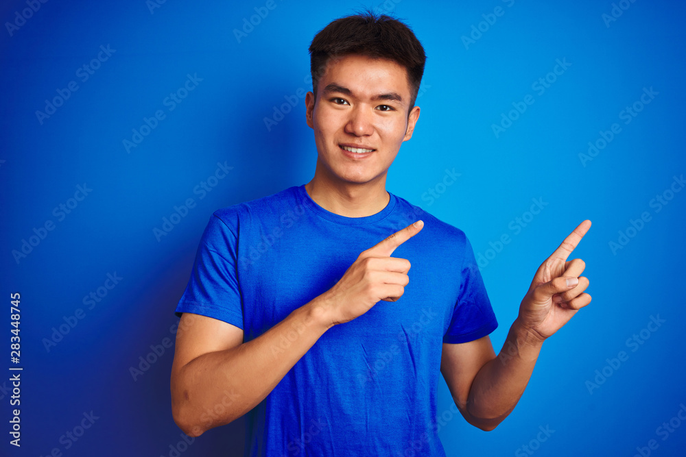 Young asian chinese man wearing t-shirt standing over isolated blue background smiling and looking at the camera pointing with two hands and fingers to the side. Young asian chinese man wearing t-shirt standing over isolated blue background smiling and looking at the camera pointing with two hands and fingers to the side.