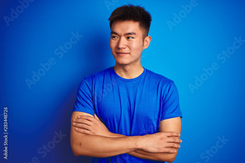 Young asian chinese man wearing t-shirt standing over isolated blue background smiling looking to the side and staring away thinking.