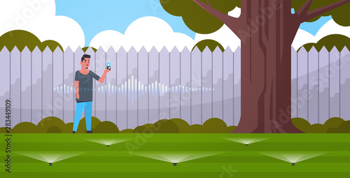 man using mobile app controlling irrigation grass watering system by smart speaker voice recognition activated digital assistant concept modern backyard with green lawn flat horizontal full length