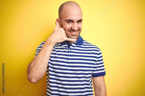 Young bald man with beard wearing casual striped blue t-shirt over yellow isolated background smiling doing phone gesture with hand and fingers like talking on the telephone. Communicating concepts.