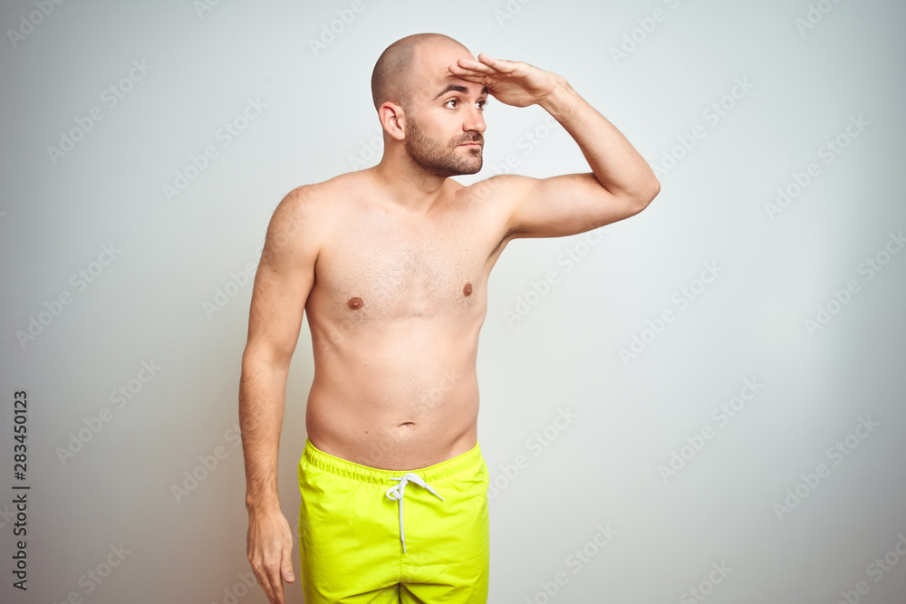 Fototapeta premium Young shirtless man on vacation wearing yellow swimwear over isolated background very happy and smiling looking far away with hand over head. Searching concept.