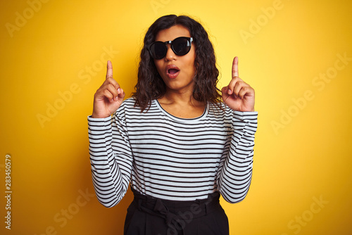 Transsexual transgender woman wearing sunglasses over isolated yellow background amazed and surprised looking up and pointing with fingers and raised arms.