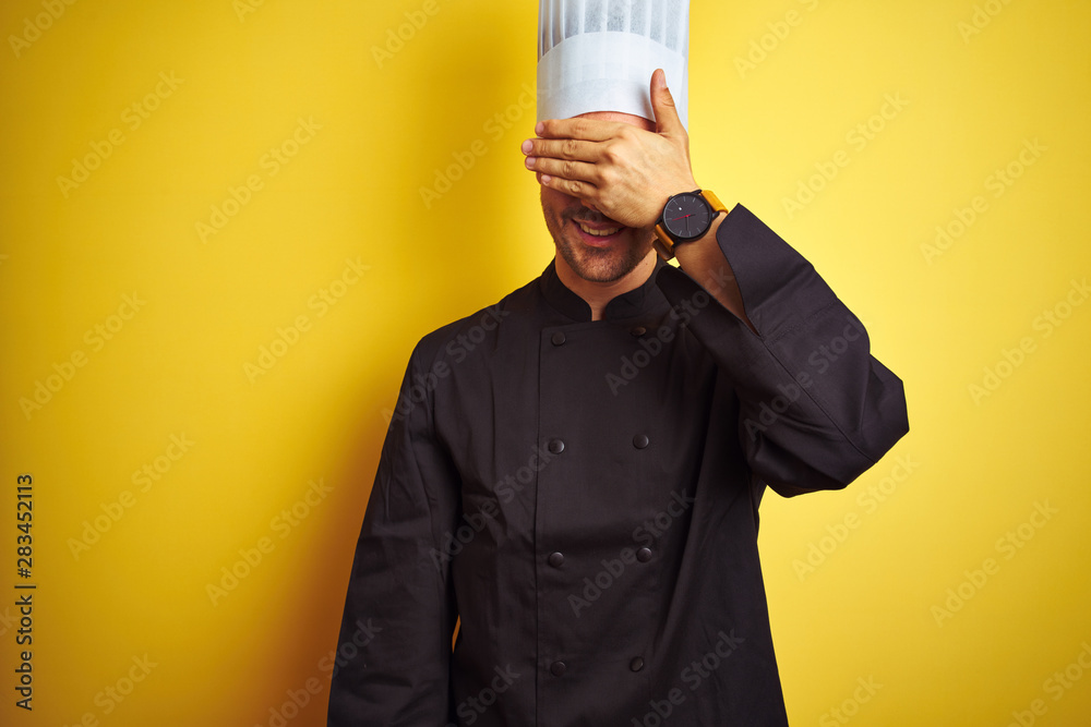 Young chef man wearing uniform and hat standing over isolated yellow background smiling and laughing with hand on face covering eyes for surprise. Blind concept. Young chef man wearing uniform and hat standing over isolated yellow background smiling and laughing with hand on face covering eyes for surprise. Blind concept.