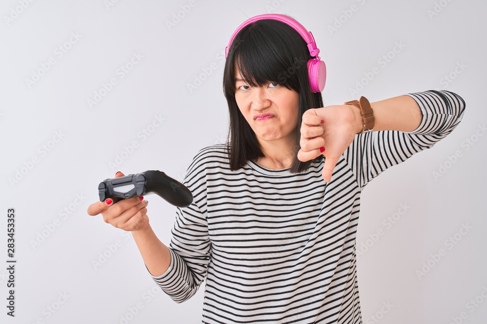 Chinese gamer woman playing video game using headphones over isolated white background with angry face, negative sign showing dislike with thumbs down, rejection concept Chinese gamer woman playing video game using headphones over isolated white background with angry face, negative sign showing dislike with thumbs down, rejection concept