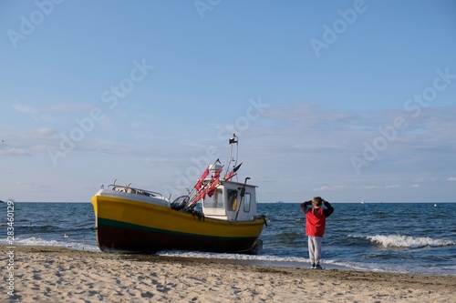 Fototapeta Naklejka Na Ścianę i Meble -  Wooden fishing boat on the beach of Baltic Sea in Sopot/Poland. Silhouette of little boy in red jacket standing near boat with raised hands and looking with delight to waves.