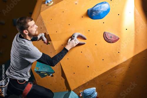 Young ambitious physically challenged man training at bouldering gym, climbing on artificial colourful rock wall, wearing harness and chalk bag, has athletic strong body.