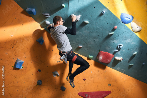 Full length shot of physically challenged boulderer with muscular body shape, trying to hold balance, hanging on holds at artificial rock wall, thinking where to place his feet.