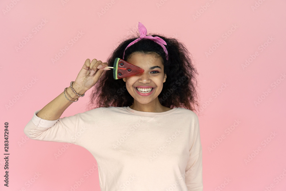 Happy smiling playful young african american teen girl black woman hold watermelon candy ice cream on face looking at camera isolated on blank pink summer studio background, portrait, copy space Happy smiling playful young african american teen girl black woman hold watermelon candy ice cream on face looking at camera isolated on blank pink summer studio background, portrait, copy space