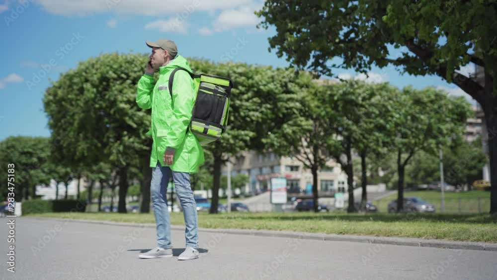 Stressed food courier walking down street having unpleasant phone call with customer or employer. Delivery guy yelling in cell phone, kicking insulated backpack, taking off uniform and walking away Stressed food courier walking down street having unpleasant phone call with customer or employer. Delivery guy yelling in cell phone, kicking insulated backpack, taking off uniform and walking away