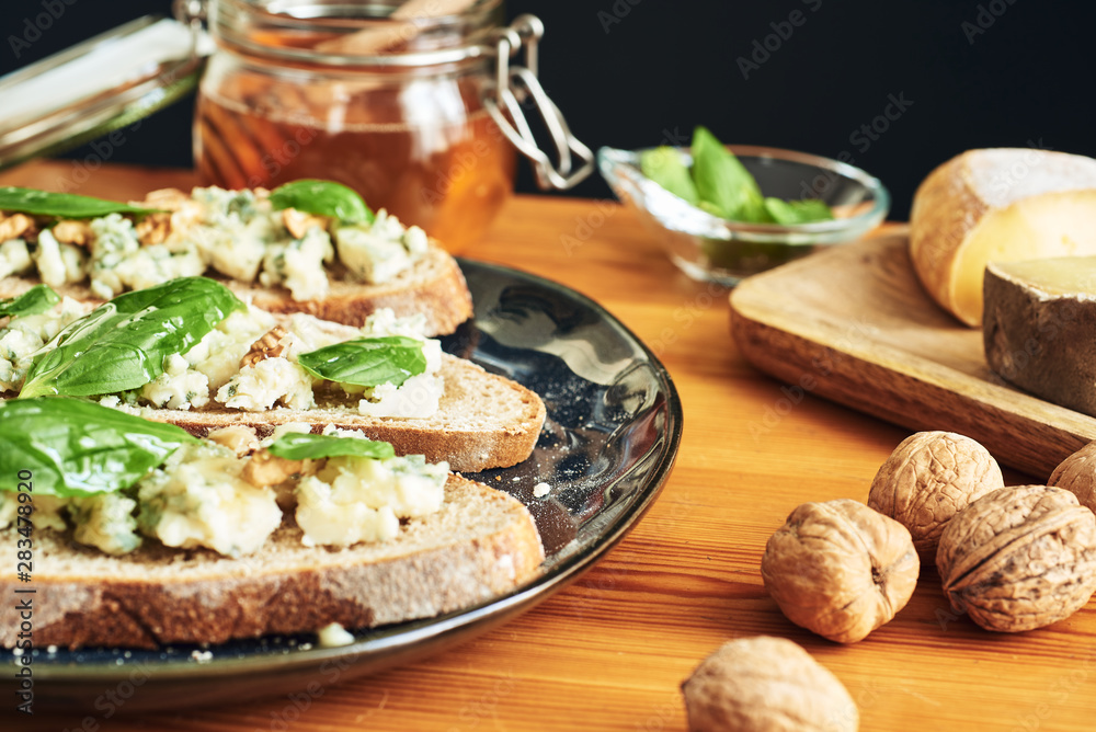Close up on a plate of toasts with roquefort cheese, walnuts, honey and basil. Some cheese, nuts and honey in the background