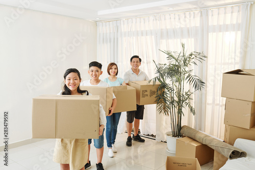Joyful Vietnamese teenagers and their parents standing in empty living room with cardboard boxes of belongings